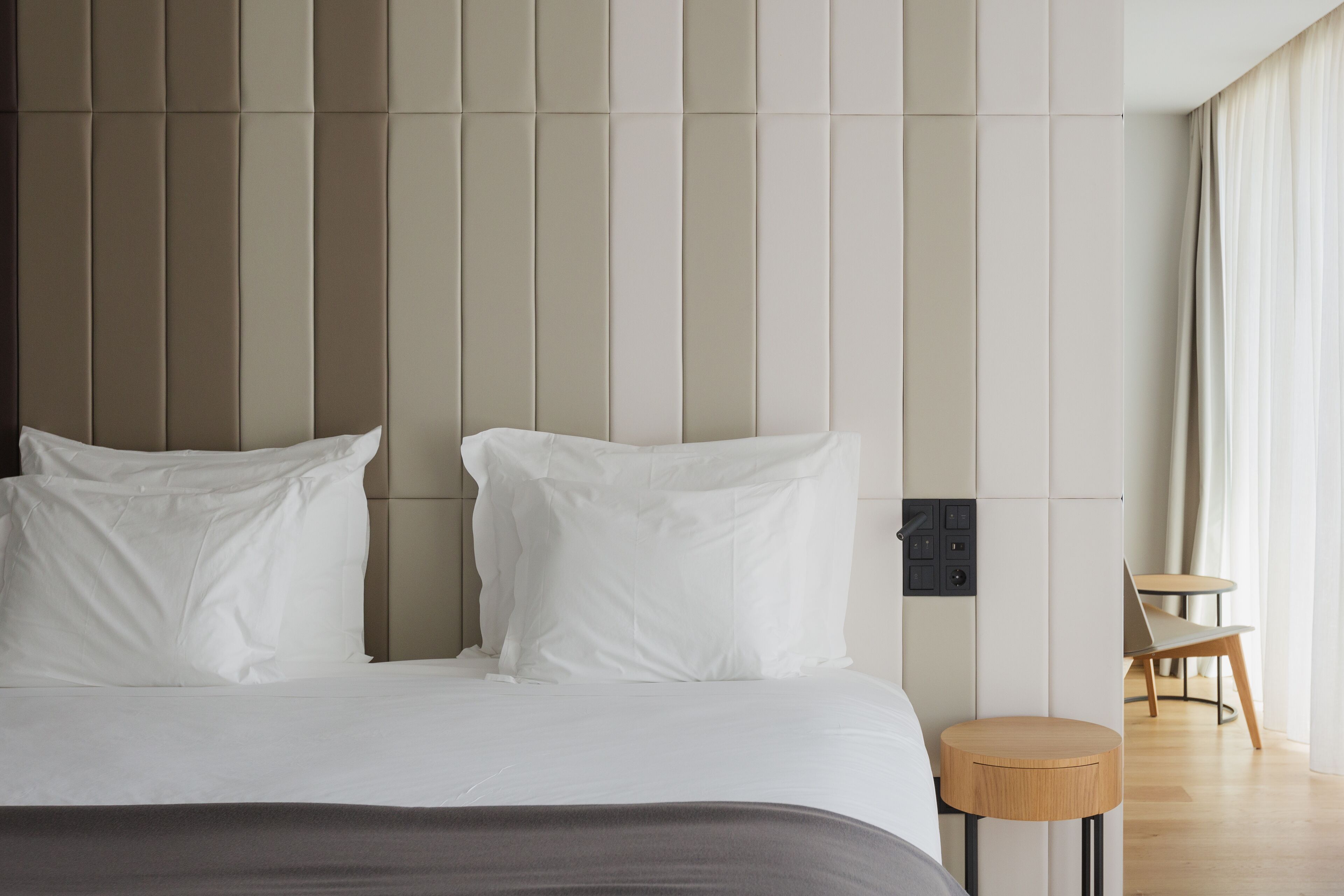 A modern hotel room with a large bed, a small wooden side table, and a view of the outdoors through a window.