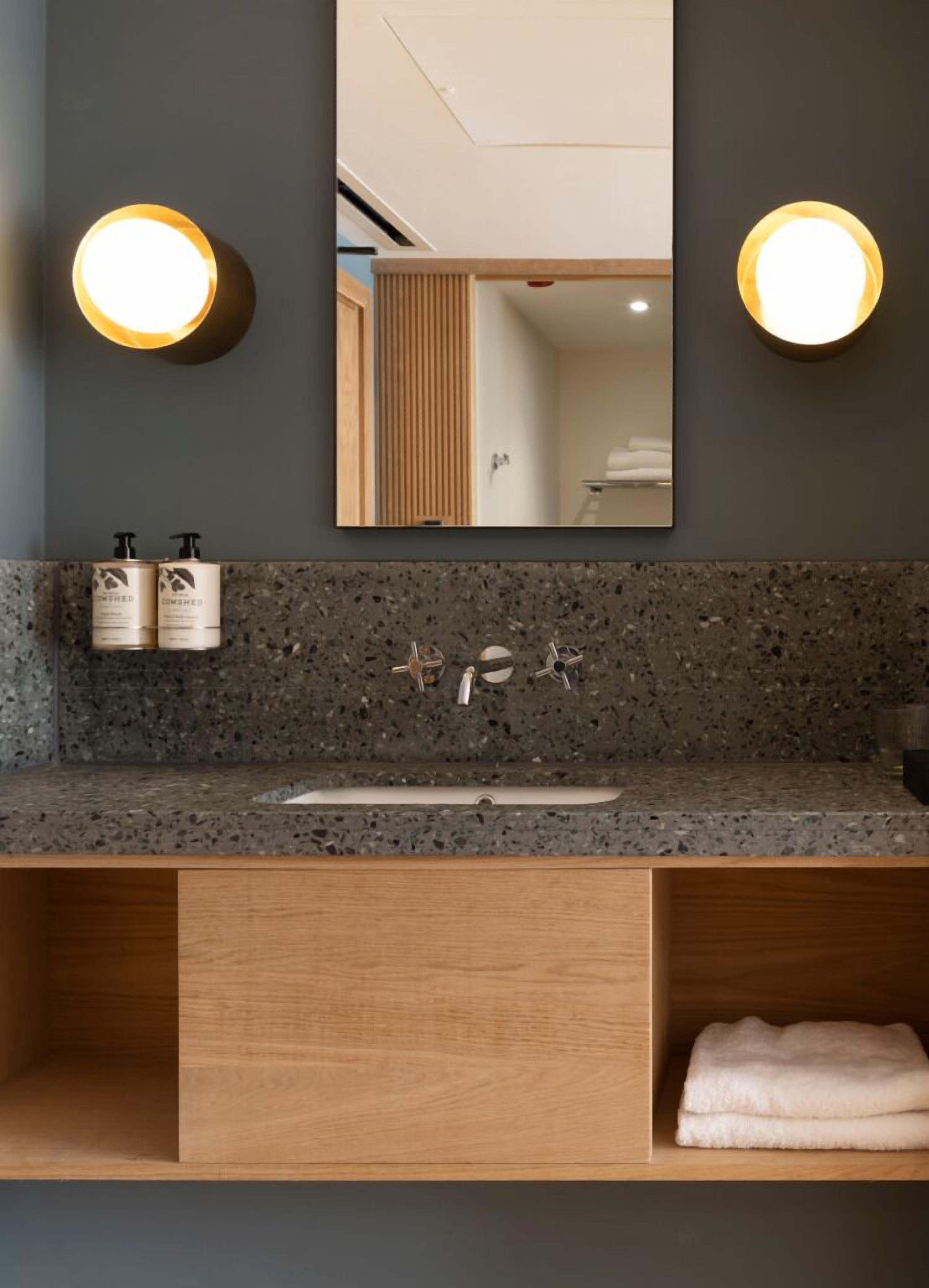 A modern bathroom with a granite countertop, a wooden vanity, a large mirror, and two wall-mounted lights.