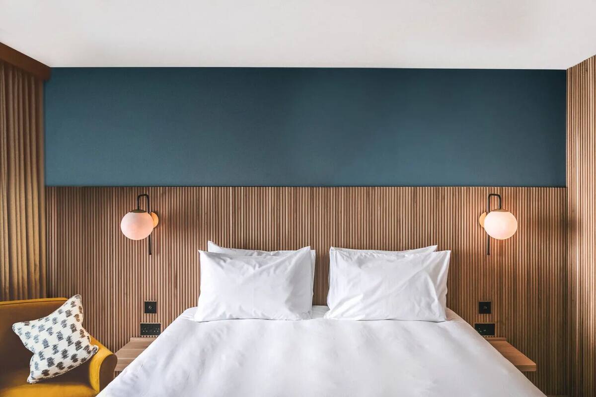 A modern hotel room with a wooden headboard, two white pillows, a bed, a nightstand with a lamp, and a patterned pillow on a yellow chair.