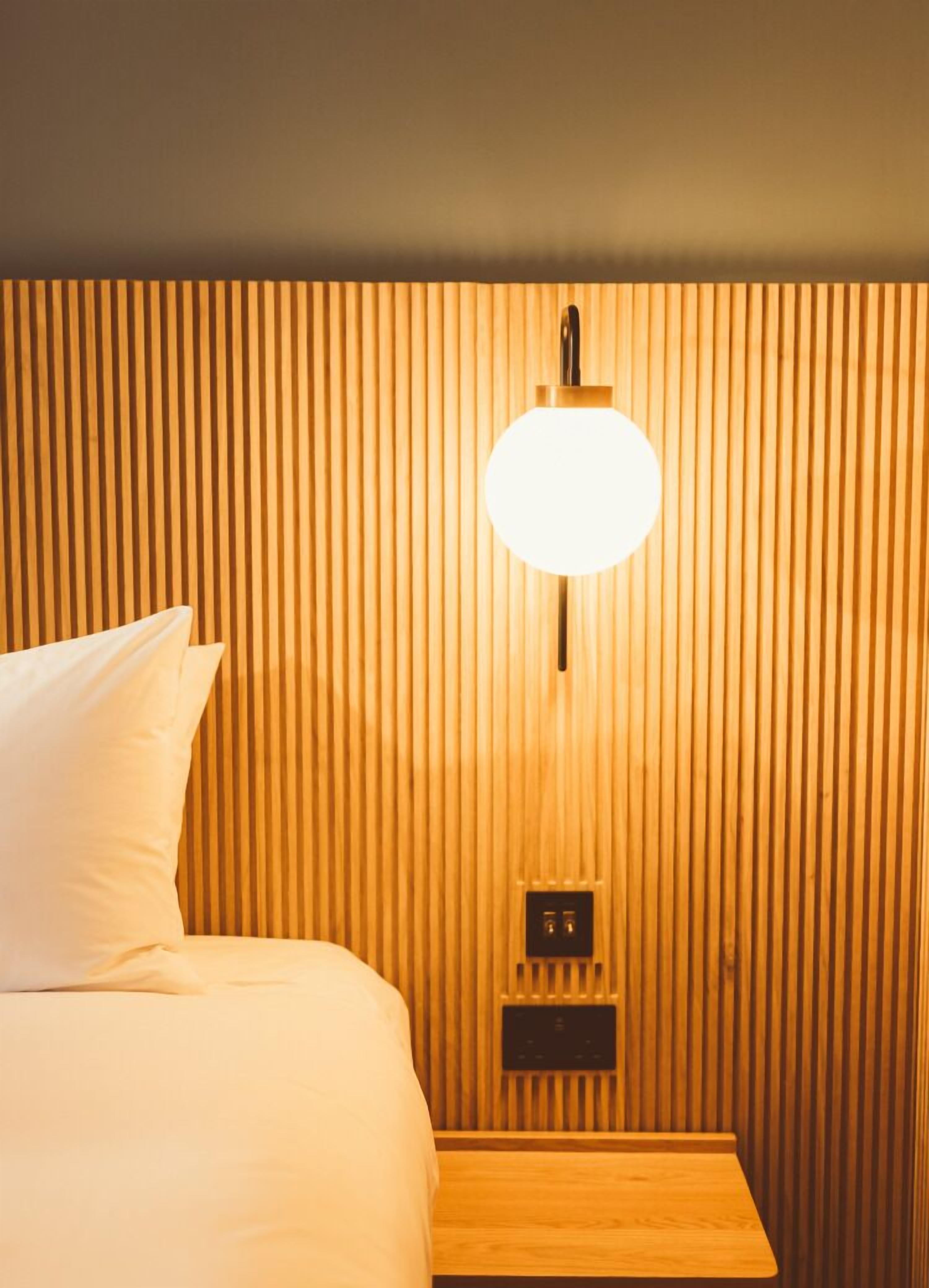 Habitación de hotel con cama, mesita de noche, lámpara de pared y panel de control.