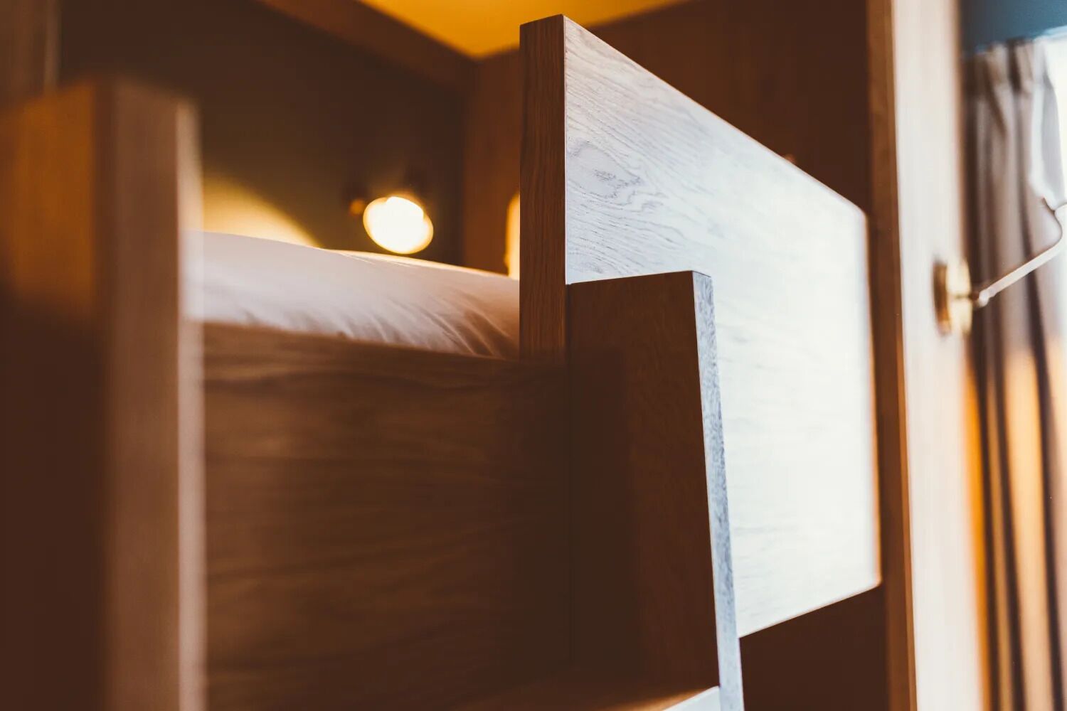 Una cama litera de madera con cabecera, una lámpara de noche y una ventana con cortinas.