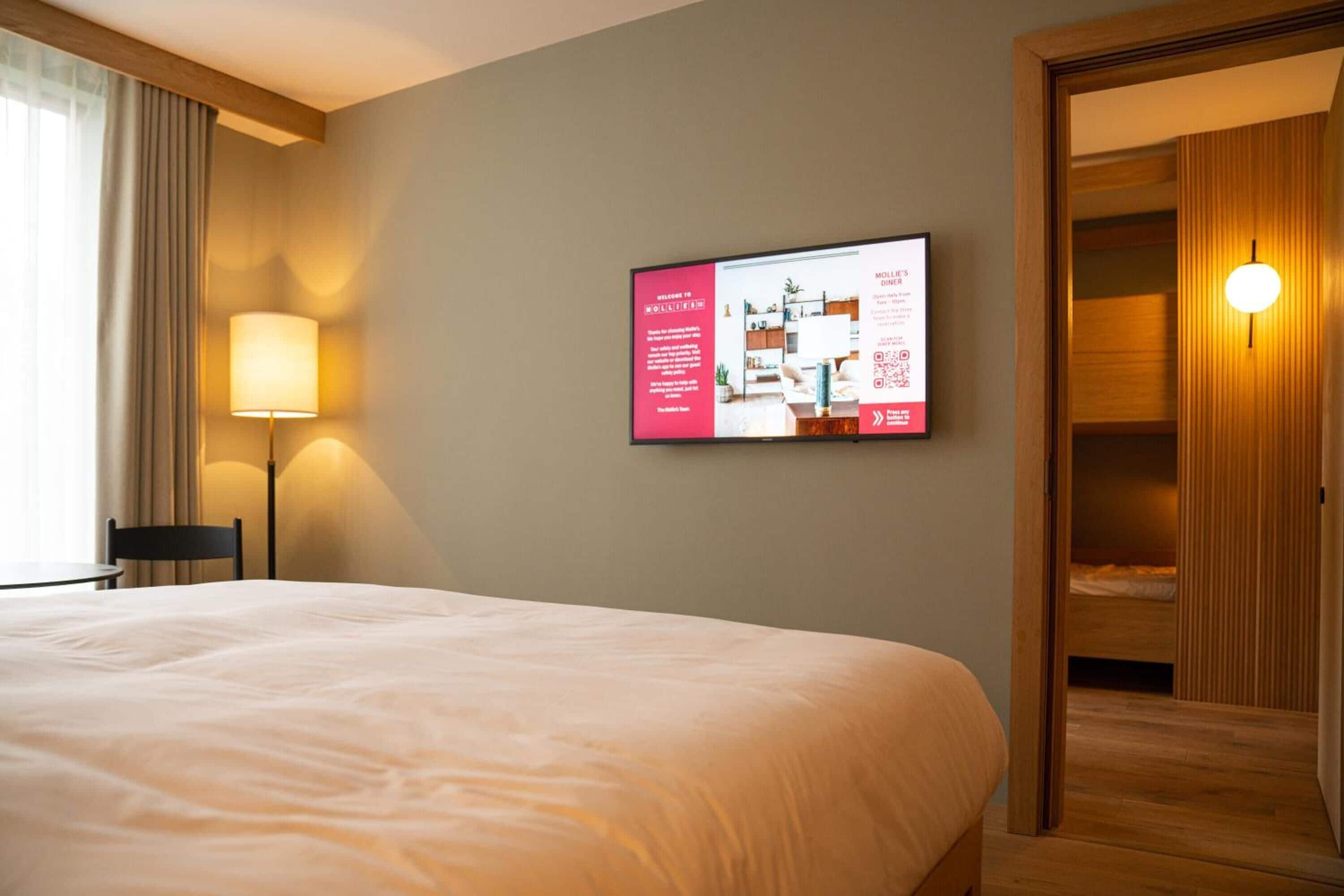 A hotel room with a large bed, a desk, a chair, a lamp, a wardrobe, and a wall-mounted screen displaying information.