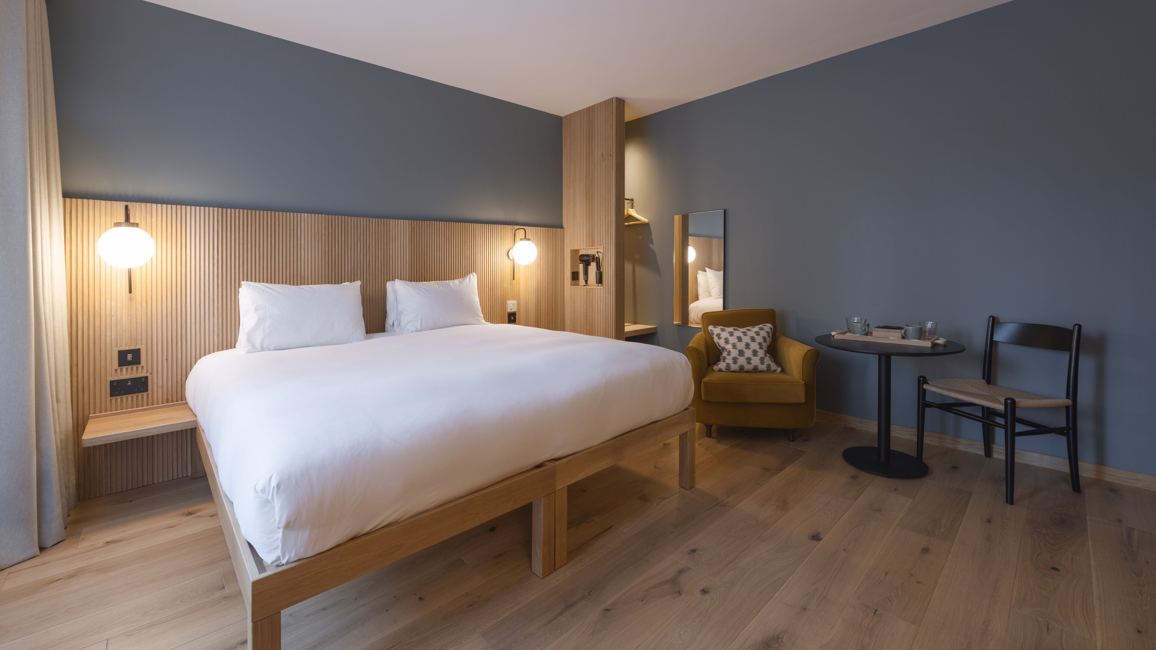 Une chambre d’hôtel moderne avec un grand lit, des tables de chevet, une chaise, une petite table et un miroir.