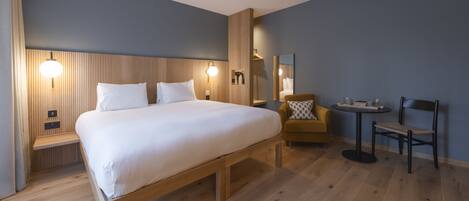 A modern hotel room with a large bed, bedside tables, a chair, a small table, and a mirror.