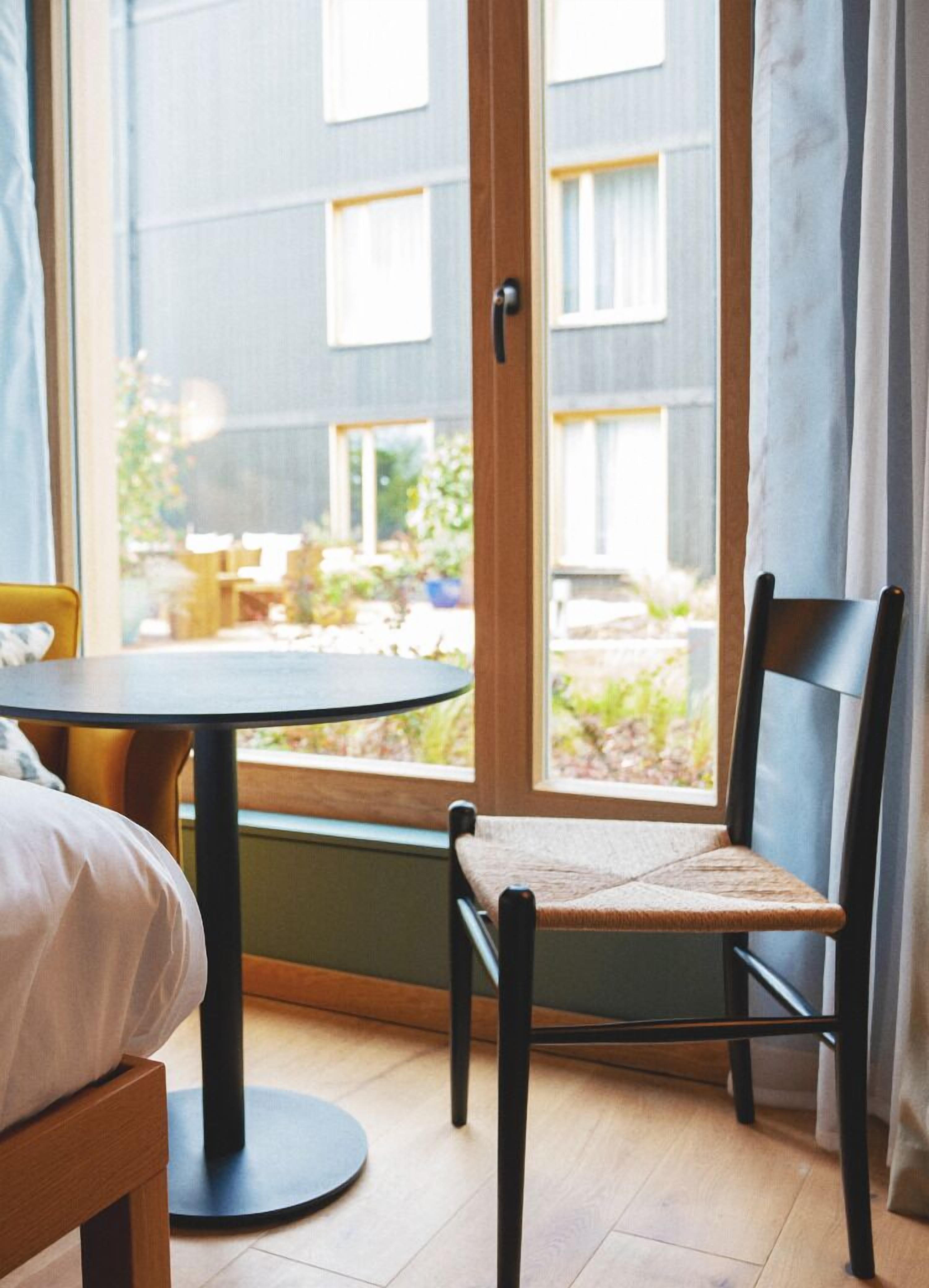 A room with a round table and a chair, a bed, and a window with curtains.