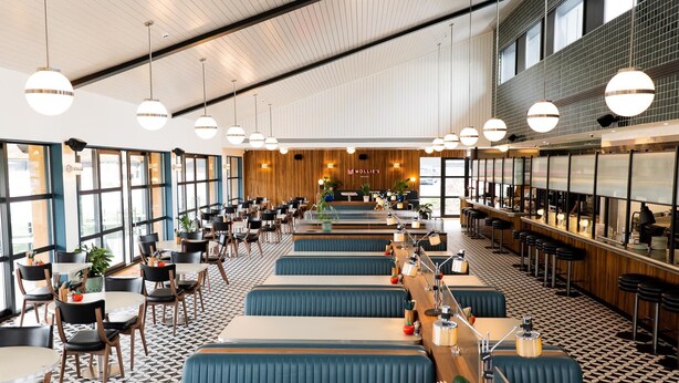 Un'ampia sala da pranzo con lunghe tavolate condivise e posti a isola, ampie finestre e lampade a sospensione.