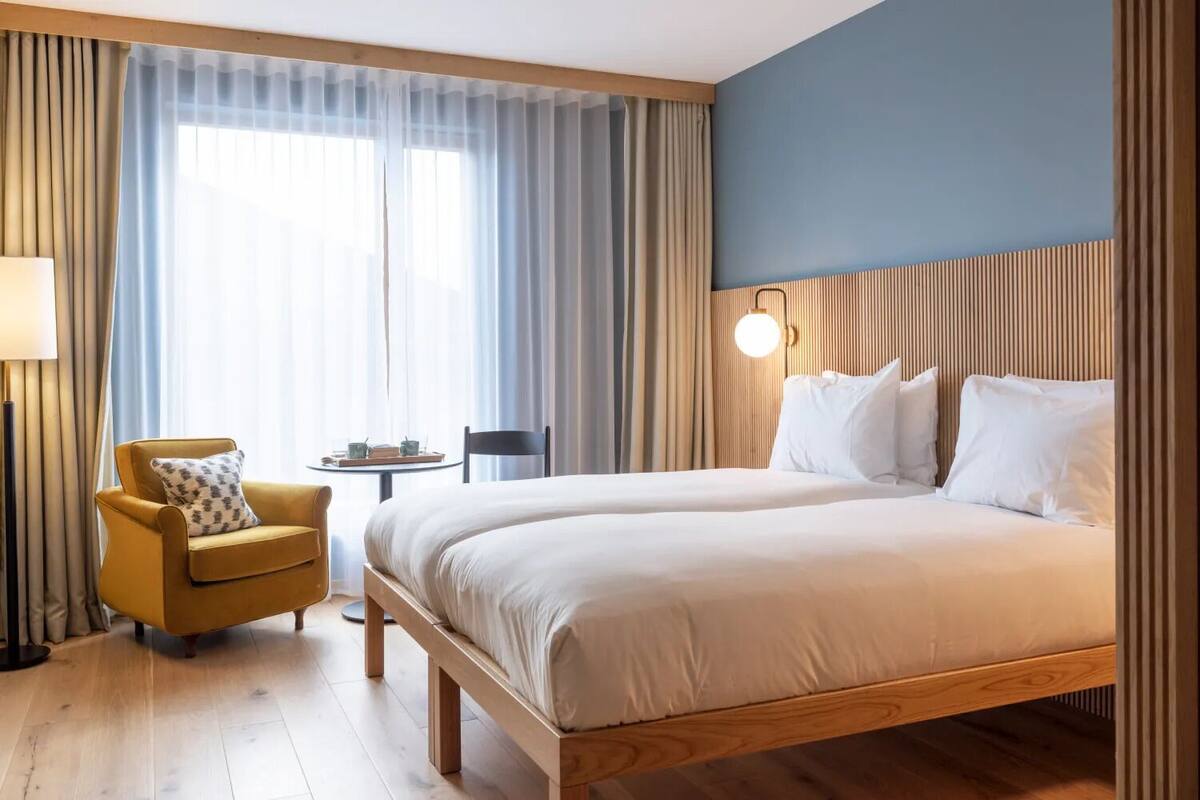 A hotel room with a large bed, a small round table with a chair, a lamp, and a window with curtains.