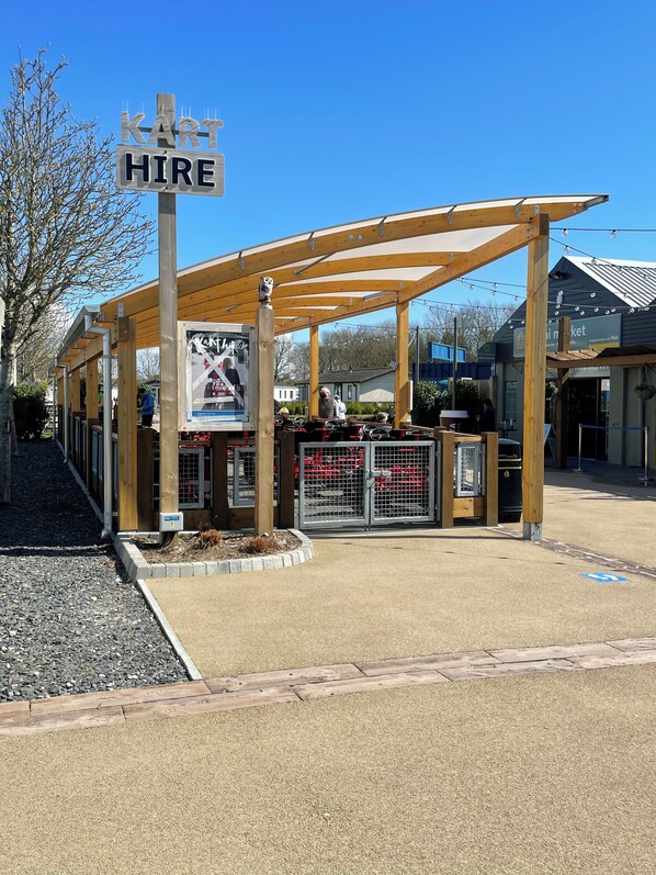 A covered outdoor seating area with a 'KART HIRE' sign, a wooden pergola, and a fenced parking area. - Beautiful 3-bed Caravan Situated on Lakeland Haven (Grange-over-Sands)