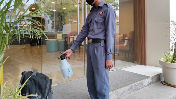 A person in uniform standing on a staircase with a spray bottle, near a glass door leading to an interior space with furniture and plants. - Kanak Beacon Hotel  (Ahmedabad)