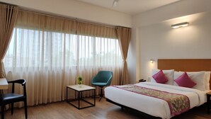A hotel room with a bed, a desk, a chair, a small table with a plant, and a ceiling fan. - Kanak Beacon Hotel  (Ahmedabad)