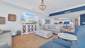 A modern living room with a grey sofa, a white coffee table, and a blue armchair. There is a flat-screen TV mounted on the wall, a chandelier, and a kitchen area visible in the background. - New to the Rental Market, Newly Owner, Newly Renovated!!! (Orange Beach)
