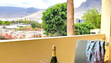 A balcony with a table, two wine glasses, and a bottle of champagne. In the background, there are mountains, trees, and a view of a town.