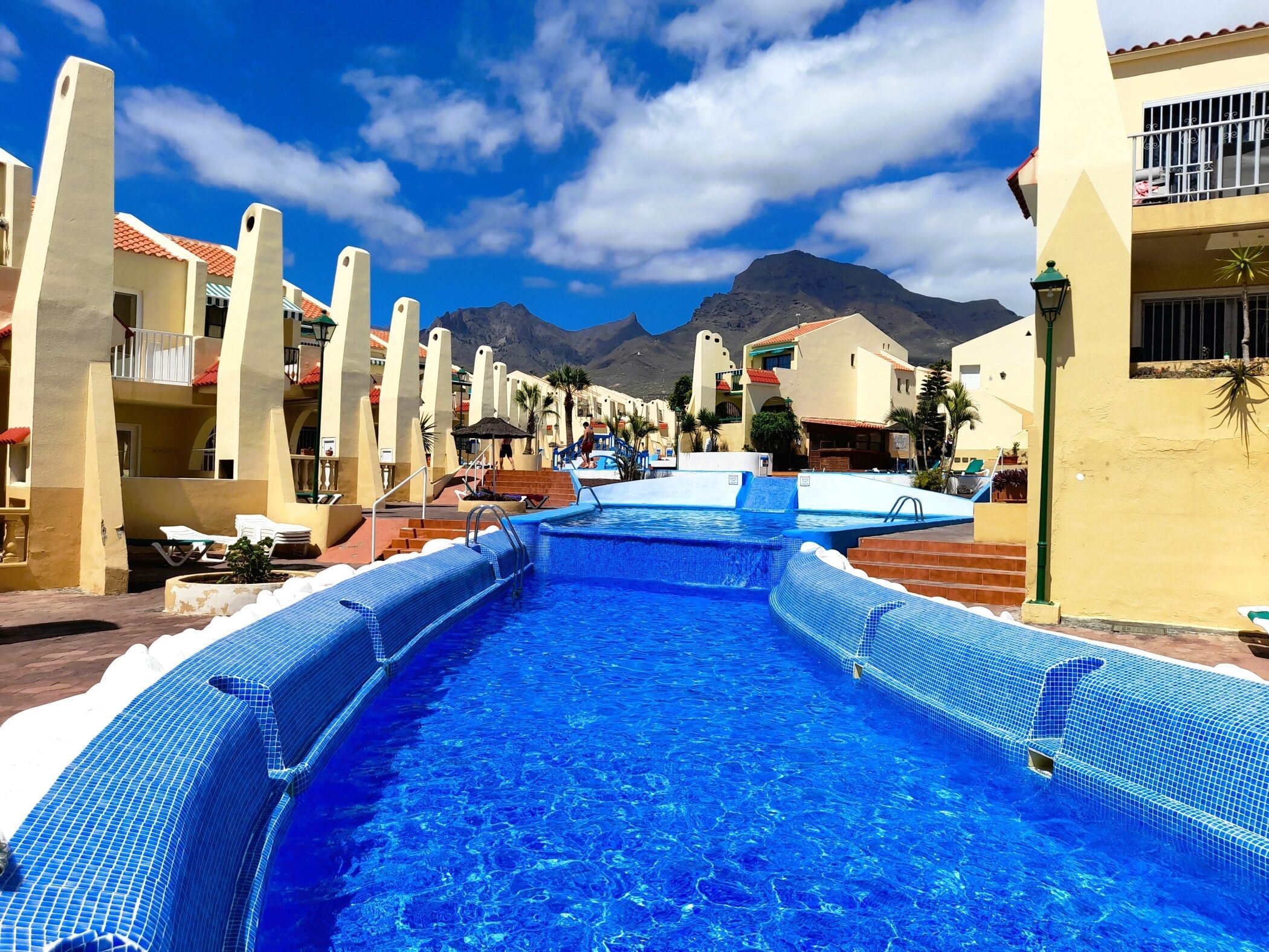 A swimming pool with a mountain backdrop, surrounded by buildings with balconies and a clear sky.