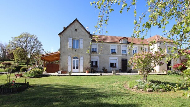 Un bâtiment à deux étages, doté d’un toit en pente, d’un porche abrité et d’un jardin bien entretenu où poussent diverses plantes et fleurs.