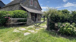 Een stenen cottage met een houten poort, omringd door weelderig groen en bloeiende planten.