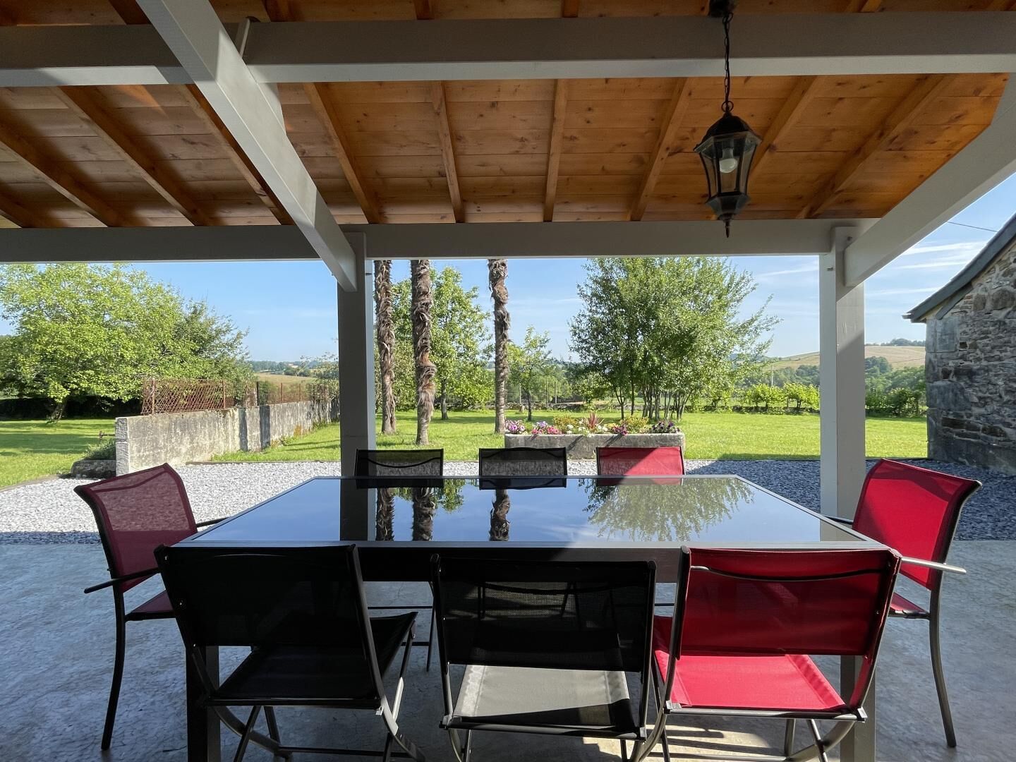 Un espace extérieur abrité, aménagé pour les repas, avec une table et des chaises, une lanterne suspendue, et une vue sur un paysage verdoyant, avec des arbres et un mur de pierres.
