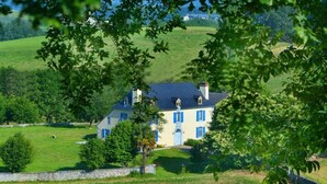 A large house with a white facade and blue shutters, surrounded by a lush green landscape and fields.