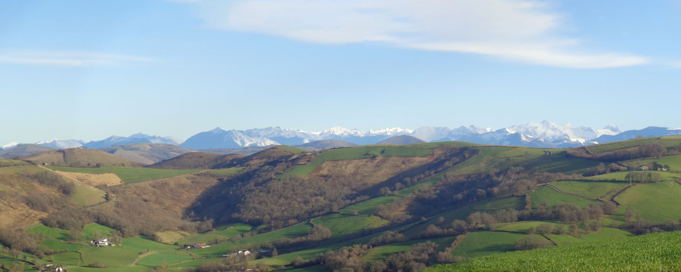 Ein weiter Panoramablick auf ein Tal mit sanften Hügeln, einer kleinen Siedlung und schneebedeckten Bergen im Hintergrund.