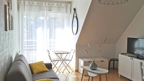 A modern living room with a grey sofa, a small white table, a TV, and a dining area with a table and chairs. - Studio Cosy au Clos Fleuri 2 Personnes (Berck)