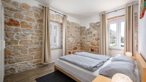 Une chambre avec un mur en pierre, un lit avec une couette à carreaux, une table de chevet en bois et une fenêtre avec des rideaux.