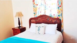 Een bed met een blauwe spreidek en twee kussens, waarvan er één een dolfijnprint heeft, in een kamer met een rieten hoofdbord en een nachtkastje met een lamp.