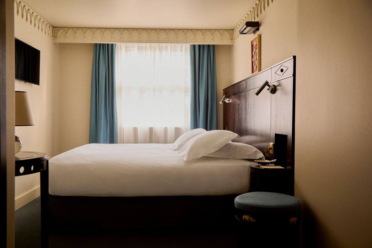 A hotel room with a bed, a nightstand, a lamp, a window with curtains, and a wall-mounted TV.