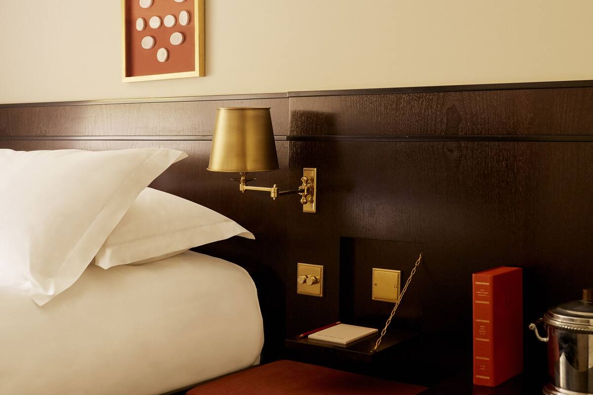 A hotel room with a neatly made bed, a bedside table with a lamp, a framed picture, and a small red box.