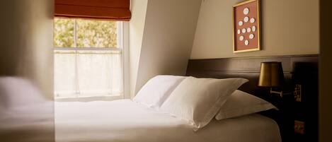 A neatly made bed with white linens in a room with a window, a framed picture on the wall, and a bedside lamp.