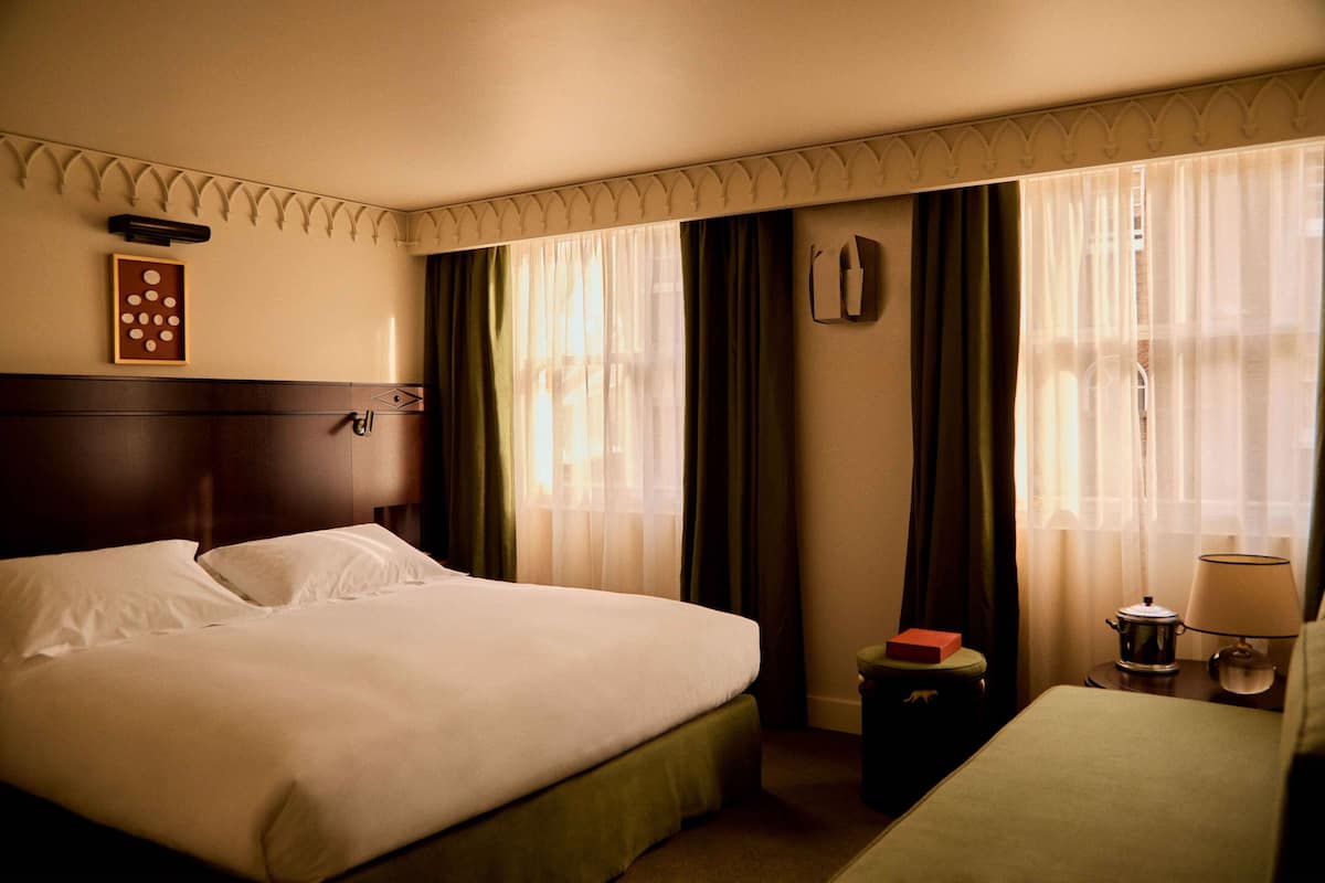 A hotel room with a large bed, a bedside table with a lamp, a small table with a red box, and a wall-mounted telephone.