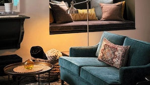 A cozy living room with a teal sofa, a round window above a built-in bench, and a small round table with a lamp. - Private Elora Studio in heart of Elora (Elora)
