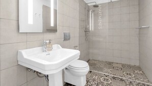 A modern bathroom with a white sink, toilet, and shower. The walls are tiled with a light color, and there's a patterned floor.