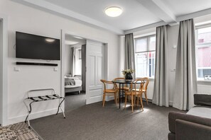 A modern hotel room with a flat-screen TV, a dining area with a table and chairs, a sofa, and a view of the outside.