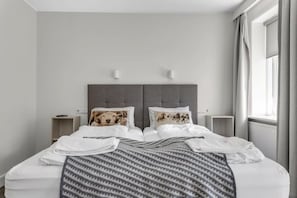 Een netjes opgemaakt bed met een gebonden deken, twee nachtkastjes met lampen, en een raam met gordijnen.
