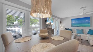 Un soggiorno moderno con zona pranzo, caratterizzato da un tavolo da pranzo in legno, sottopiatti intrecciati, un divano bianco con cuscini blu e un grande quadro raffigurante una spiaggia.