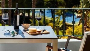 Une table dressée avec des verres à vin, une bouteille, du fromage et un saladier, offrant une vue imprenable sur un paysage luxuriant, avec des palmiers et la mer à l’horizon.