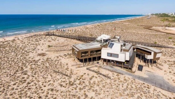 Une propriété située en bord de mer, dotée d’une structure moderne comprenant une grande terrasse et un espace couvert, et jouxtant une plage de sable et l’océan.