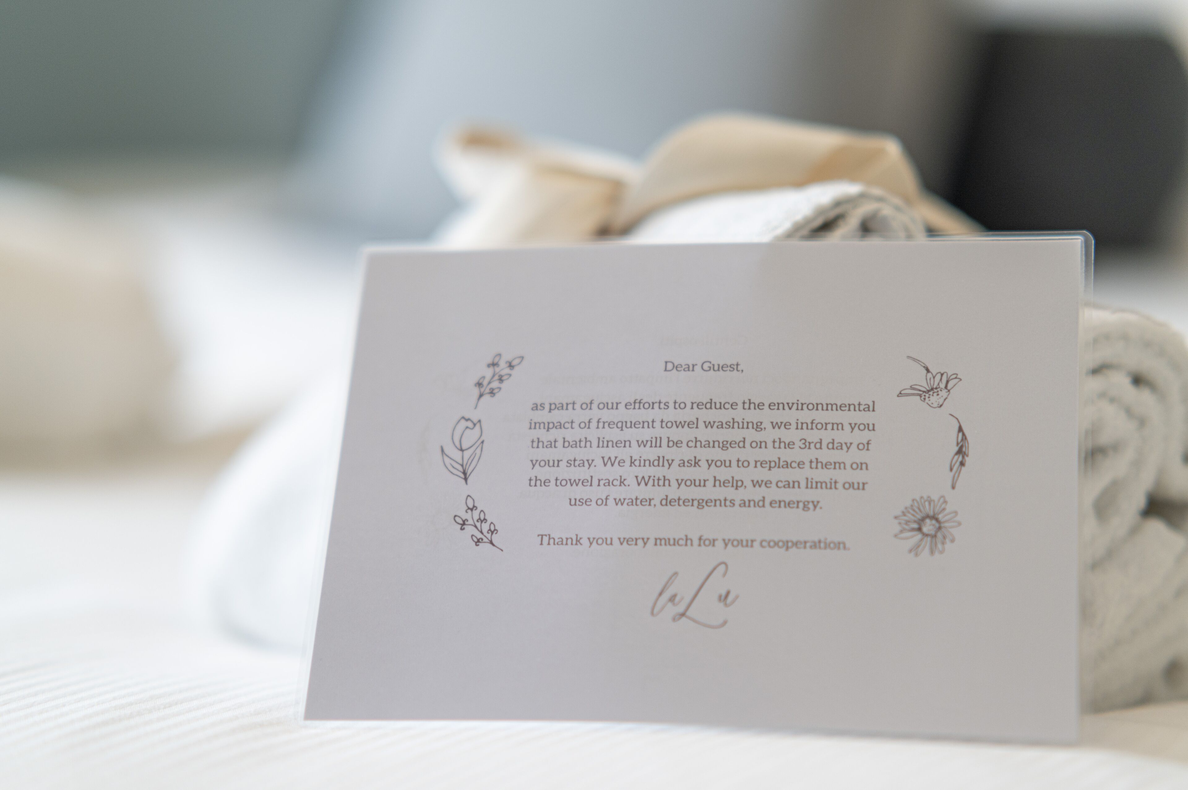 Une carte de remerciement posée sur un lit, à côté d’une serviette enroulée.