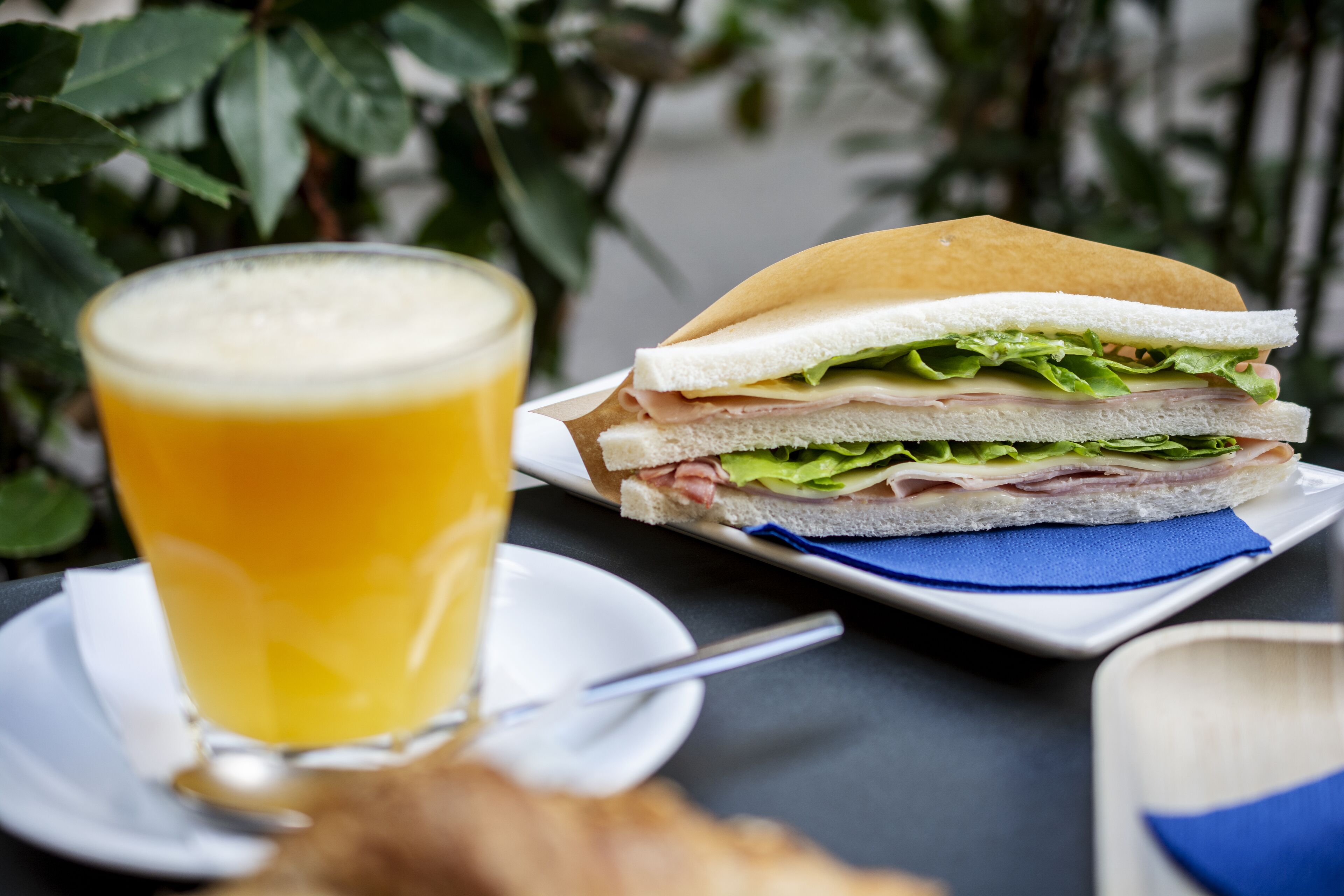 Un sándwich sobre un plato blanco con una servilleta azul, un vaso de jugo de naranja y una cuchara en un platito blanco.