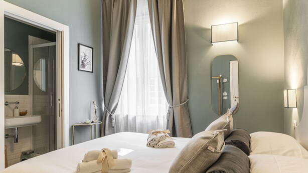 A hotel room with a bed, a bathroom with a sink, a mirror, and a window with curtains. - La Lu Cozy Rooms 2 (Pisa)