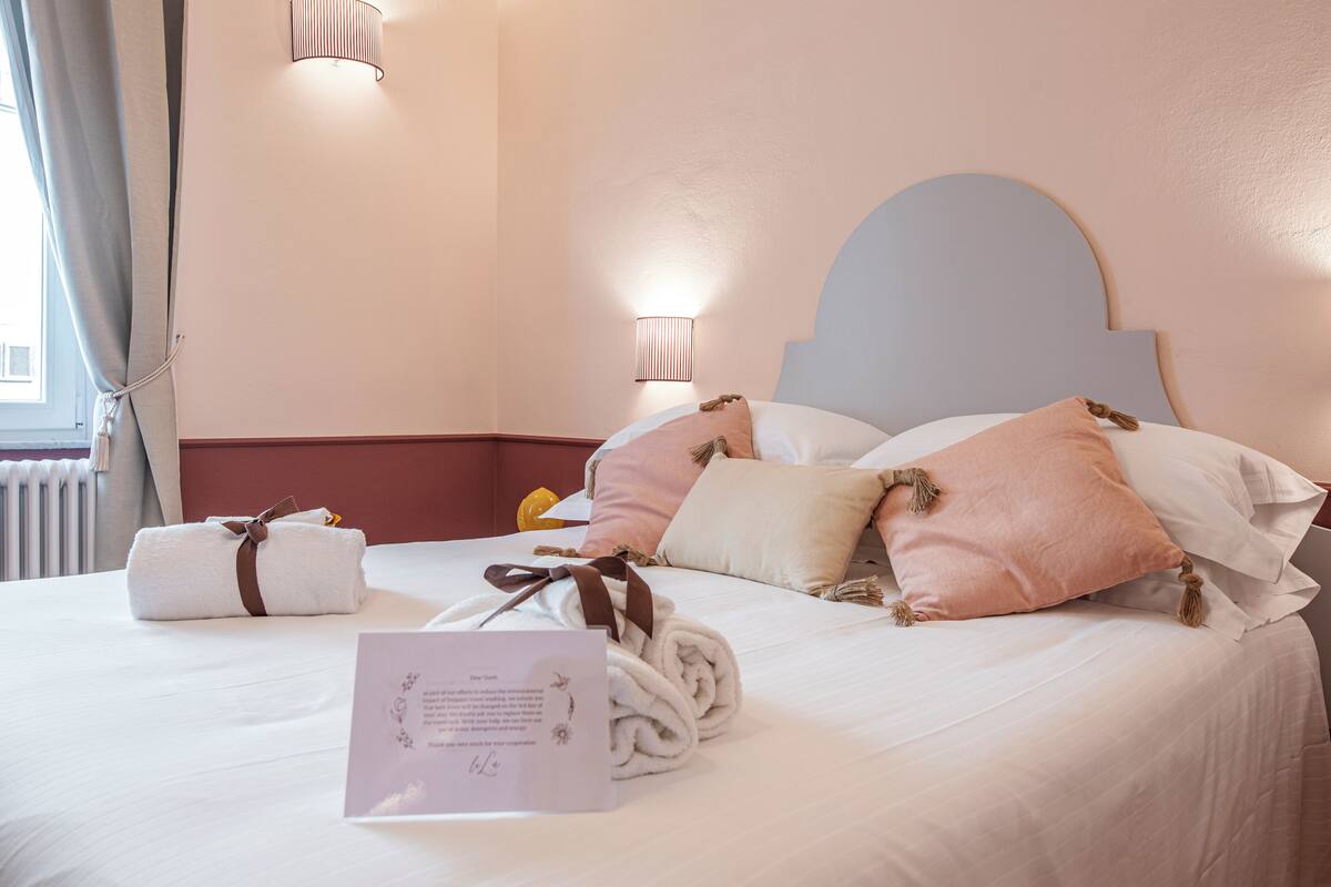 A neatly made bed with rolled towels and a welcome card, a nightstand with a lamp, and a window with curtains.