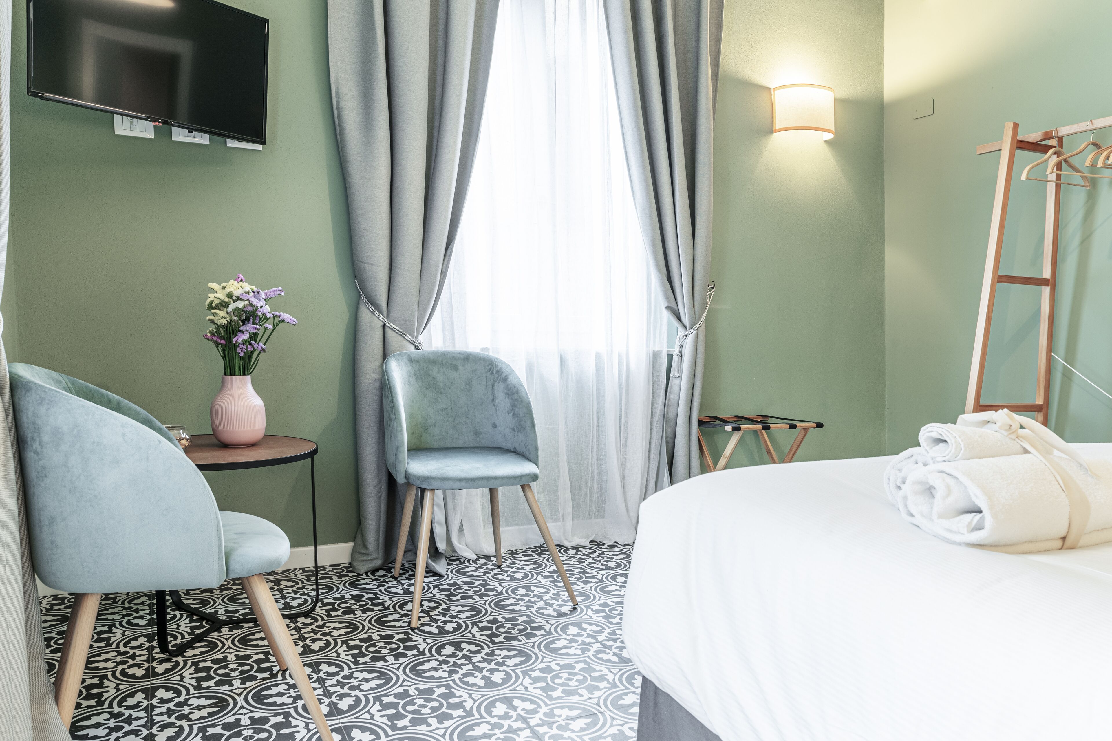 Una habitación de hotel con una cama, una silla, una mesita con un florero y una televisión instalada en la pared, además de un ventanal con cortinas.