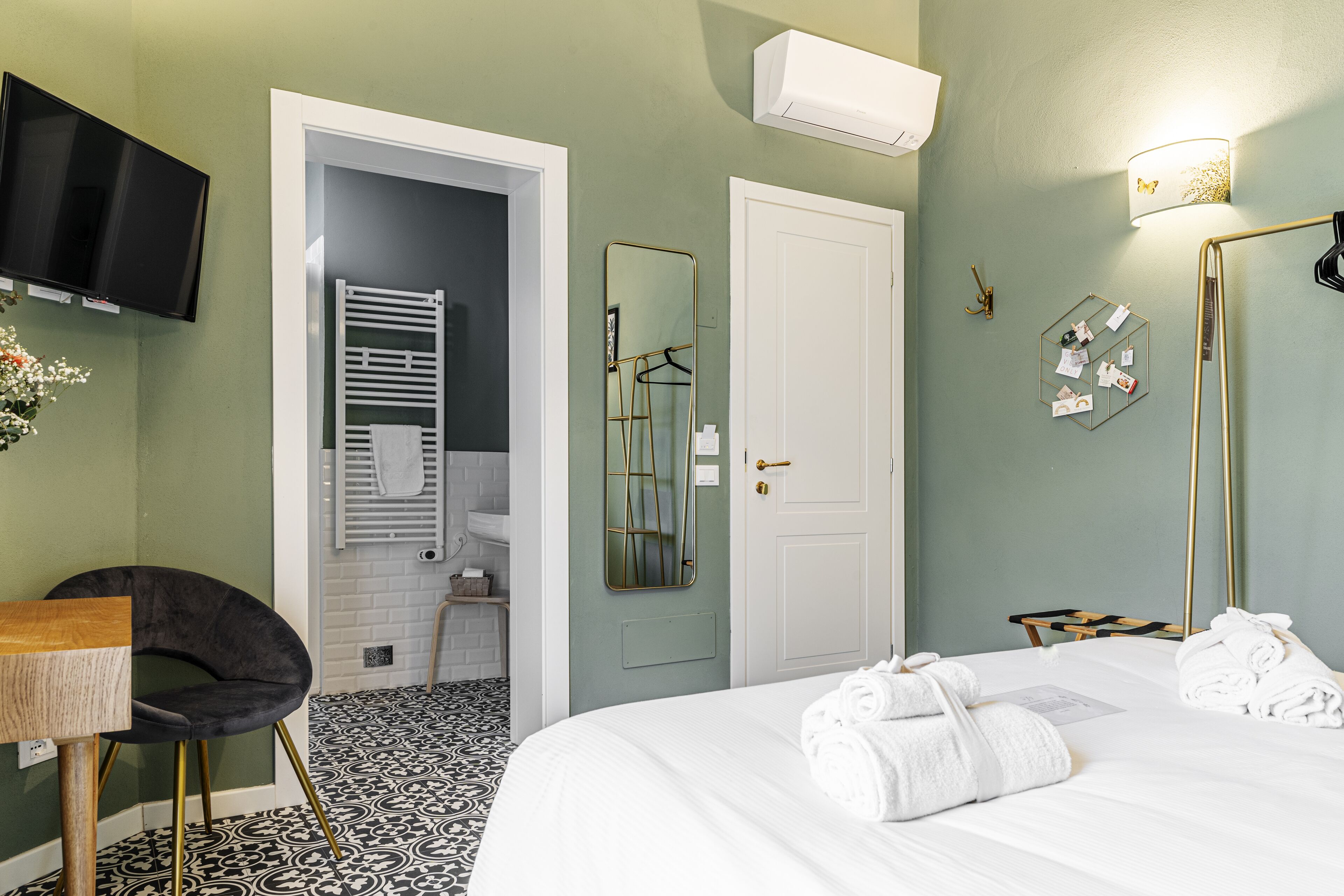 A hotel room with a bed, a TV, a chair, a mirror, and a bathroom visible through an open door.