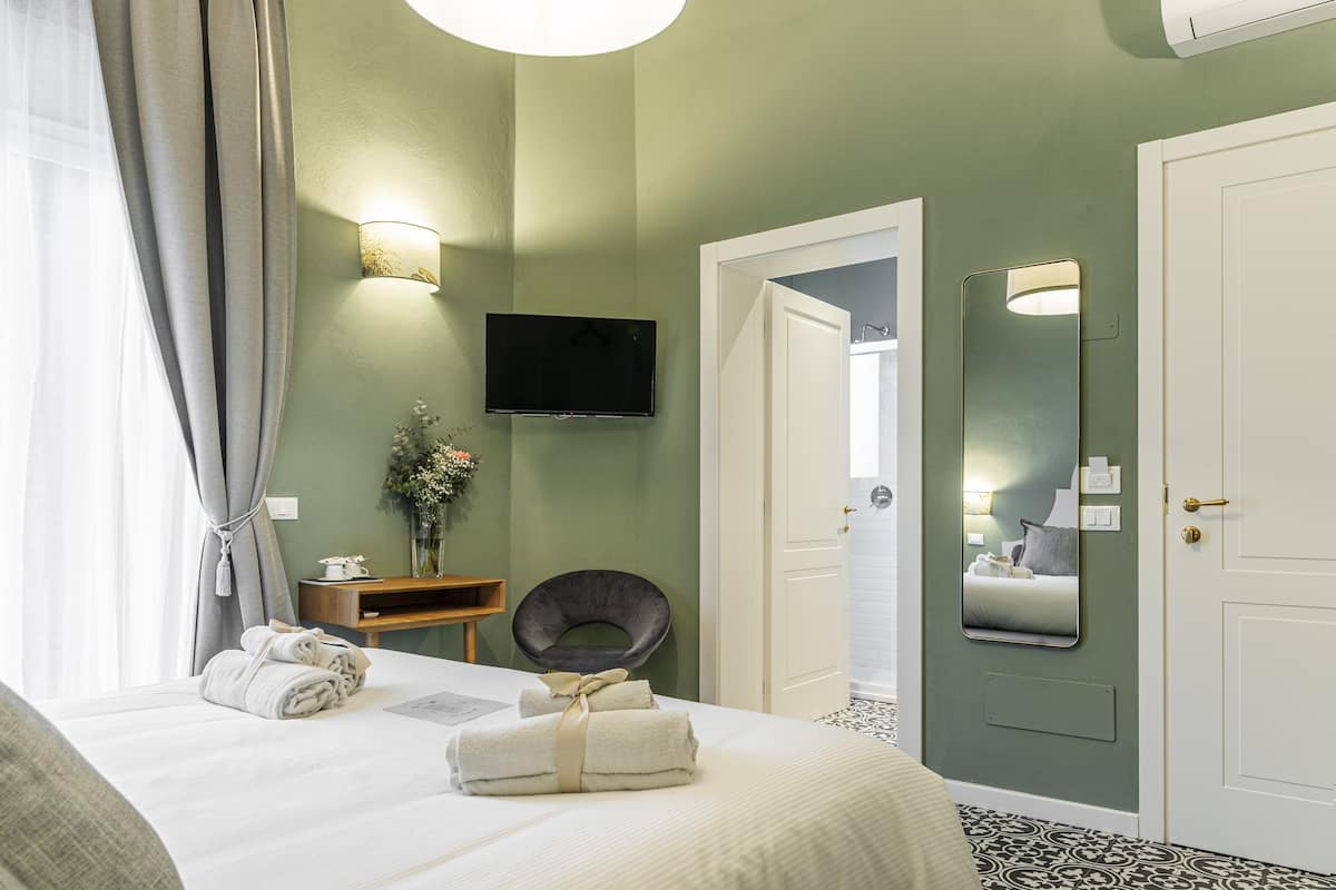 A hotel room with a bed, a flat-screen TV, a mirror, a chair, and a door leading to a bathroom.