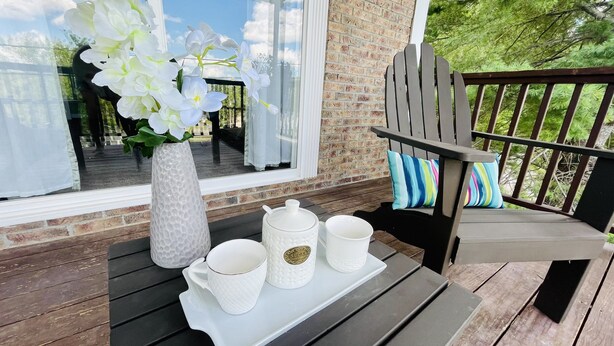 Een houten terras met een witte tafel, daarop witte keramische kopjes en een suikerpotje. Links een witte vaas met bloemen, en rechts een kussen met blauwe en groene strepen.