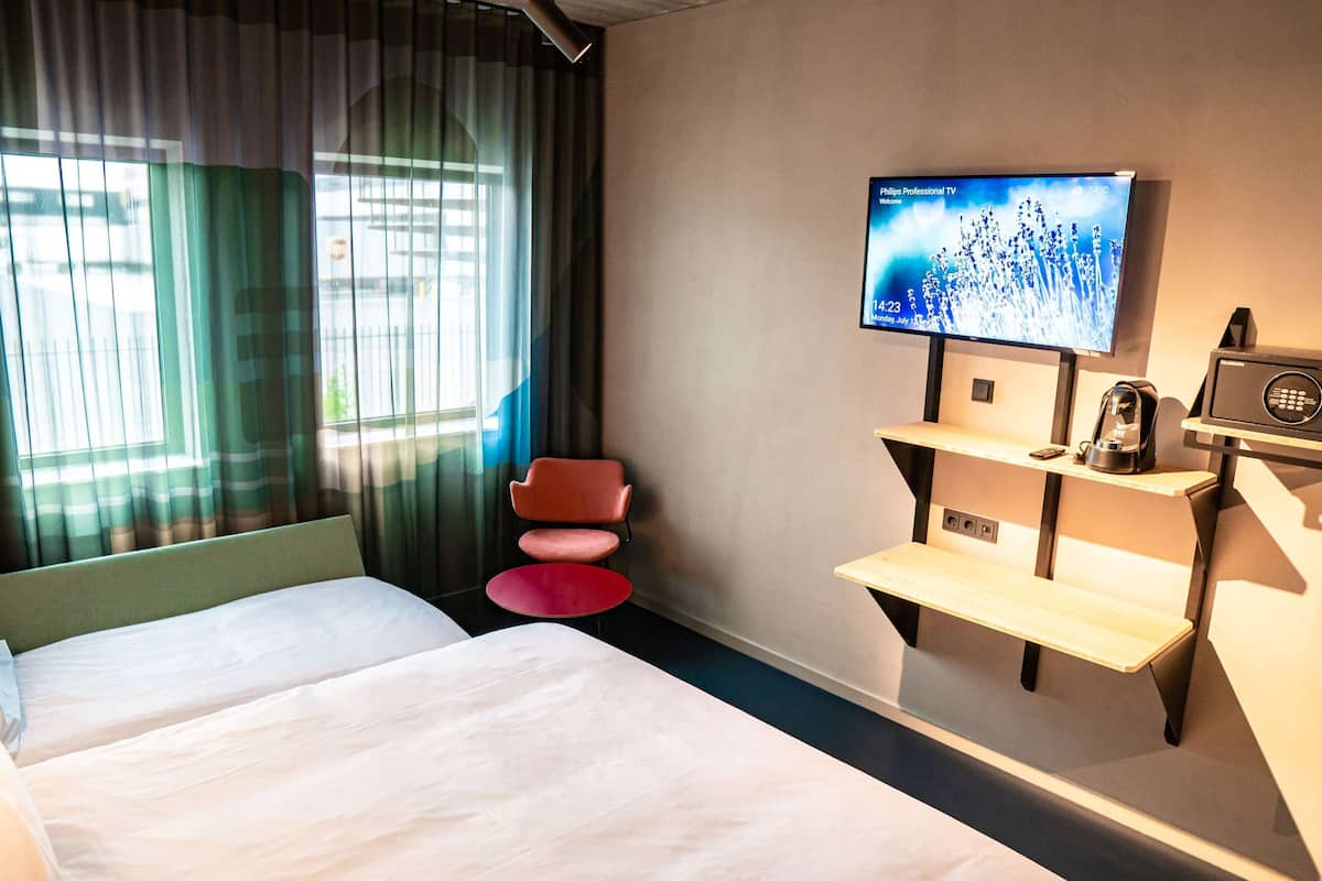 A hotel room with a bed, a TV mounted on the wall, a small table, a chair, and a window with curtains.