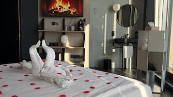 A modern hotel room with a large bed, a flat-screen TV displaying a fireplace, a bathroom with a mirror, and a view of the city.