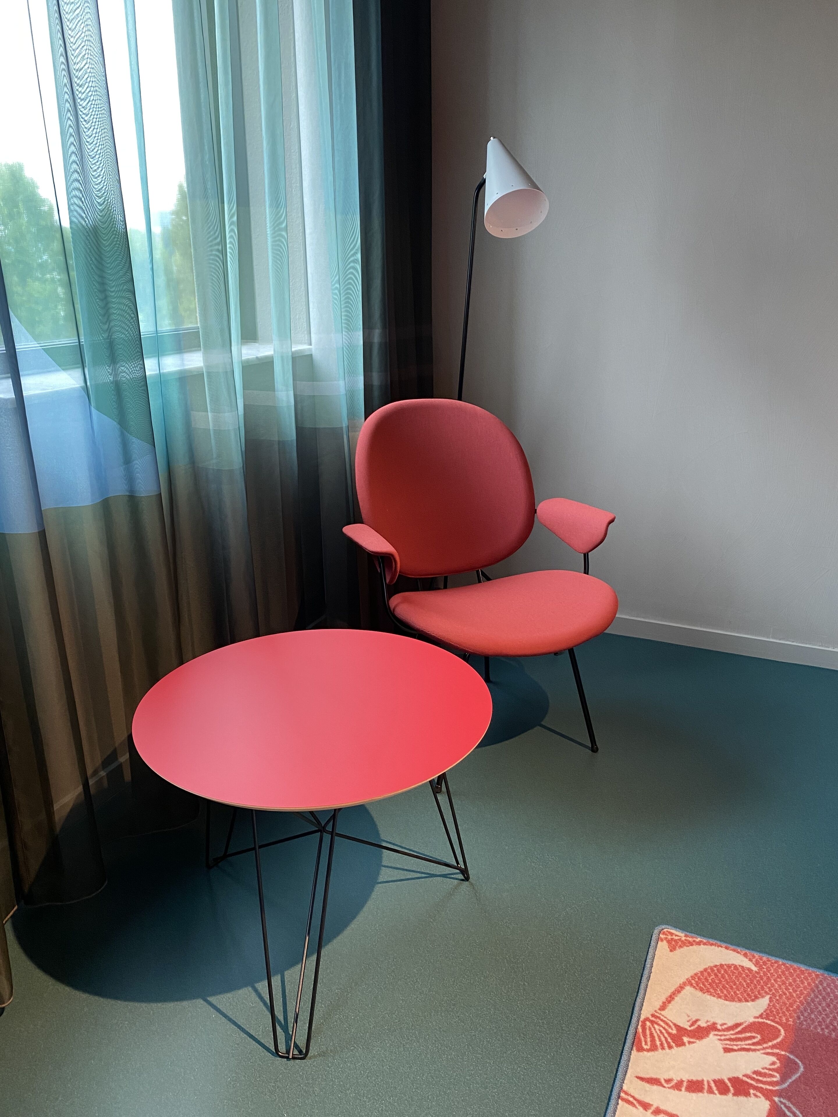 A room with a red chair and table, a floor lamp, and sheer curtains.