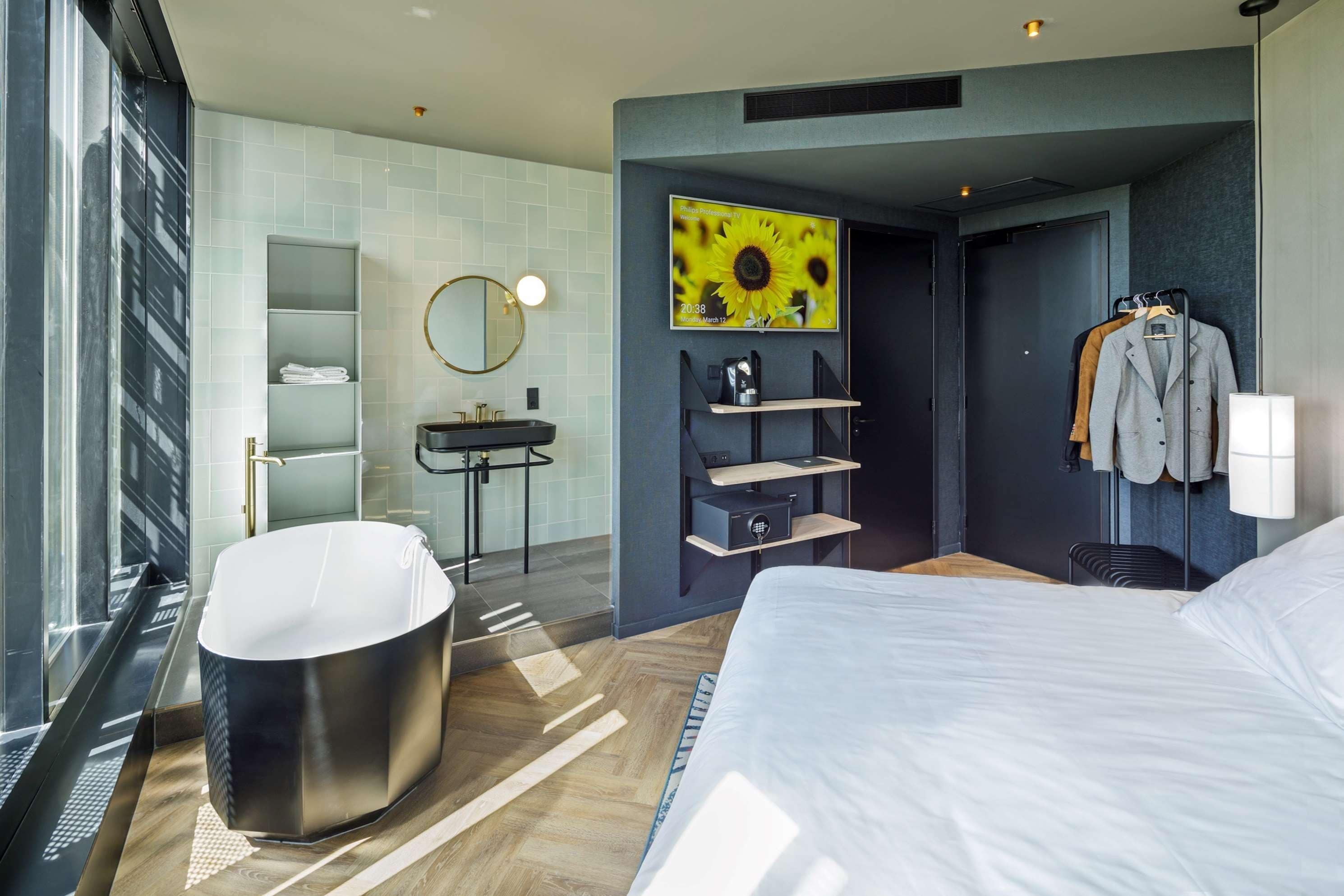 A modern hotel room with a large bed, a round mirror, a built-in sink, a shower, and a TV displaying a sunflower.