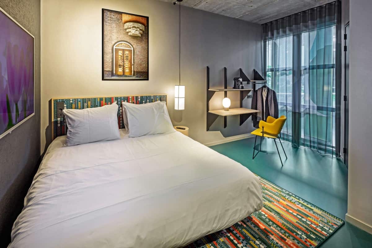 A hotel room with a large bed, a small table with a lamp, a chair, and a view of the city through large windows.