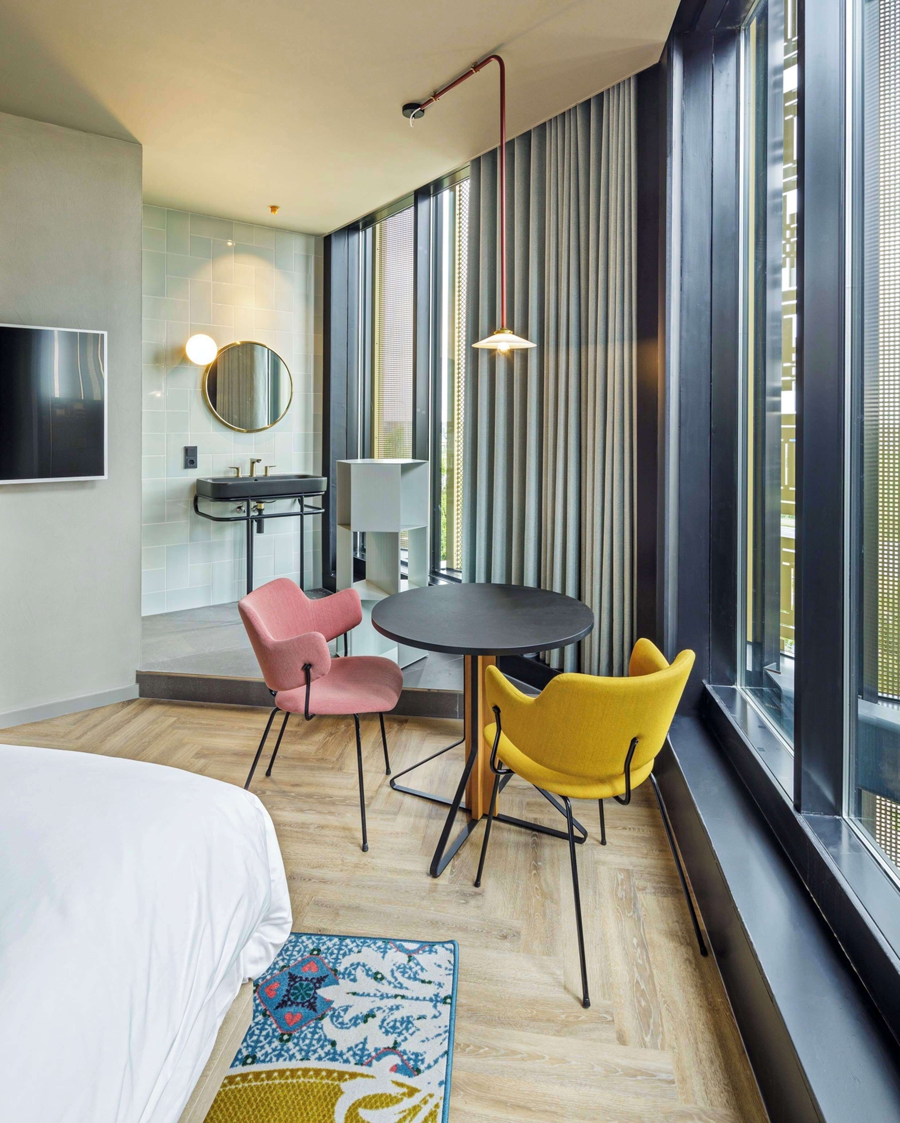 Una habitación de hotel moderna con una cama, una mesa redonda, dos sillas, un escritorio, un espejo y ventanas grandes con persianas.